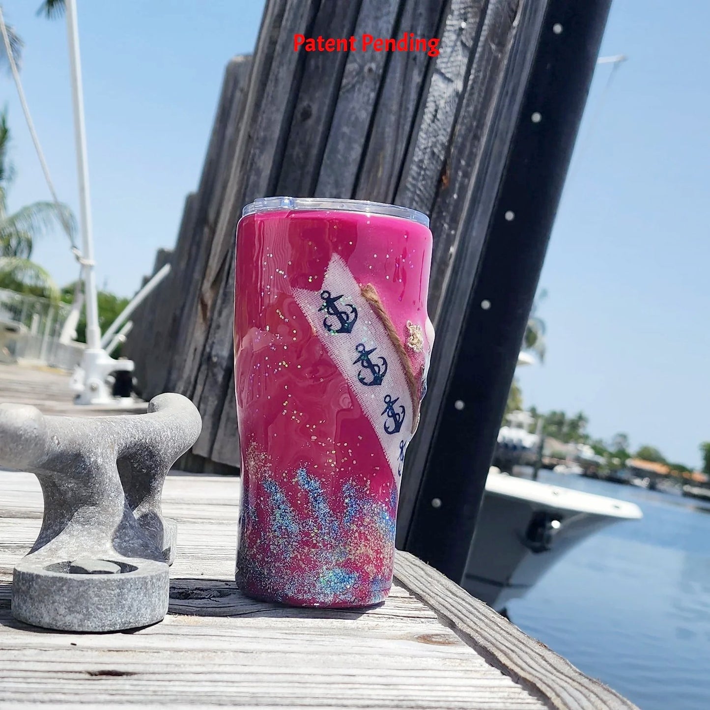 Left side view of the 20oz Saltwater Sips beach tumbler on a wooden dock, showing the detailed diagonal wrap of the anchor-print ribbon and embedded shoreline elements against the magenta color.