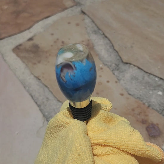 Video demonstration showing a close-up, rotating view of the smooth oval beach sand and blue ocean resin bottle stopper to display its glassy polish and natural embedded elements.