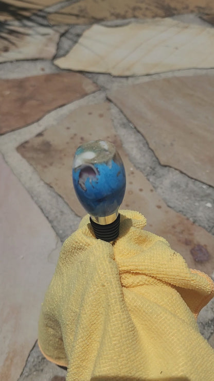 Video demonstration showing a close-up, rotating view of the smooth oval beach sand and blue ocean resin bottle stopper to display its glassy polish and natural embedded elements.