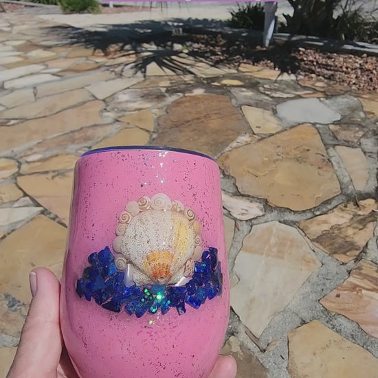 Video of a handmade pink 12oz seashell wine tumbler with lapis lazuli chips held in hand outdoors by the water