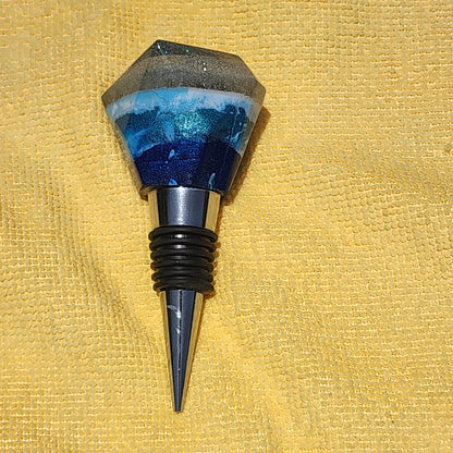 Artisan ocean-themed resin bottle stopper with distinct deep blue, aqua, and sandy beach layers, displayed on a yellow textured microfiber cloth.