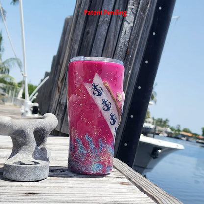 Left side view of the 20oz Saltwater Sips beach tumbler on a wooden dock, showing the detailed diagonal wrap of the anchor-print ribbon and embedded shoreline elements against the magenta color.