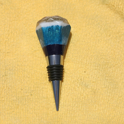 Close-up of a handcrafted geometric resin bottle stopper showcasing shimmering turquoise glitter and wave-like white and blue layers on a polished alloy stem