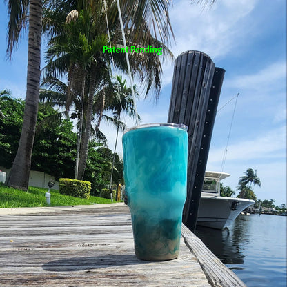 Handmade 30oz teal Caribbean coastal side view of fishing tumbler with real fish hooks, glow-in-the-dark shrimp lures, silver crab and starfish charms, and sea snail shells - Hook & Tumble™ by SoFlo Boho Boutique, displayed on a South Florida marina dock with palm trees and water in the background.