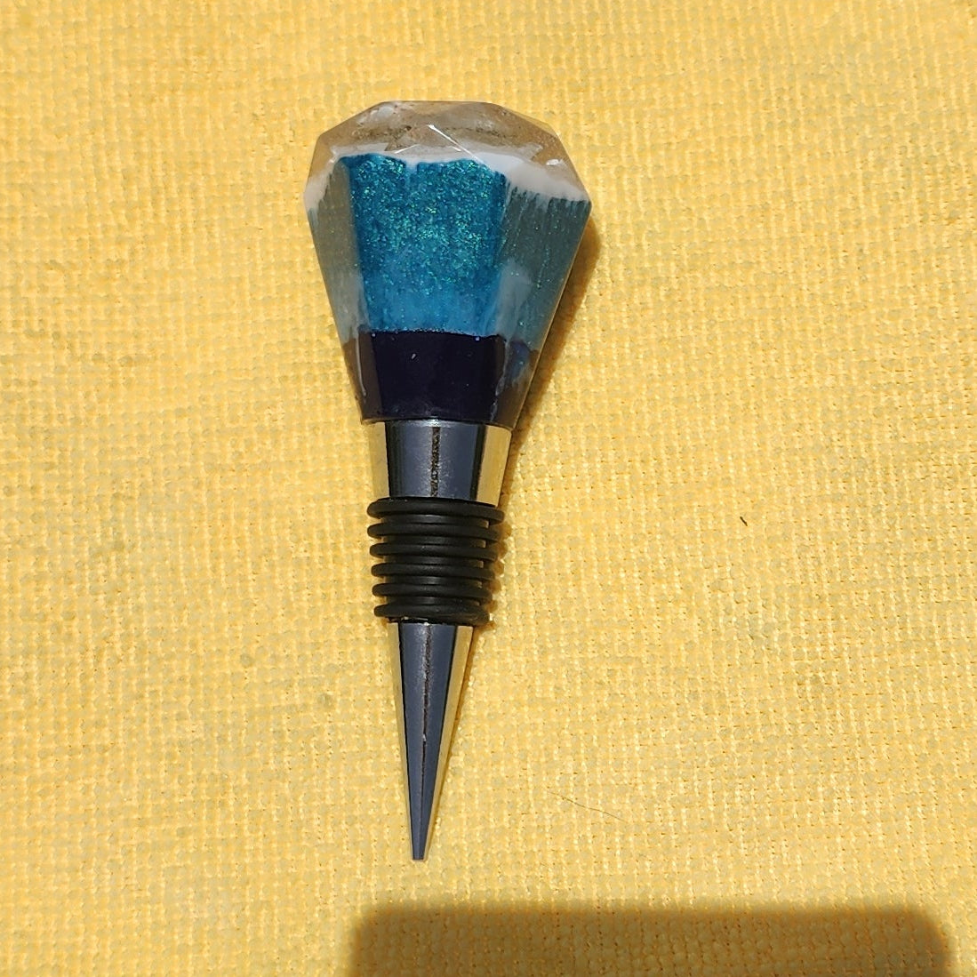 Artisan resin bottle stopper featuring a faceted teal and navy blue ocean-inspired top resting on a yellow microfiber background