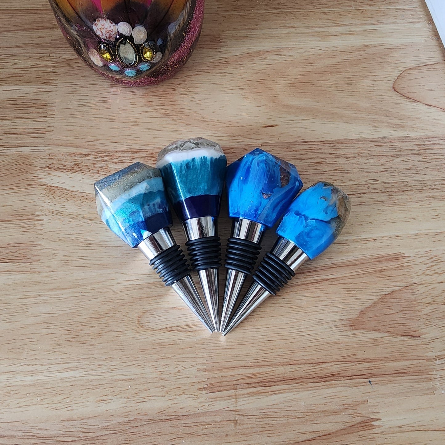 Top-down view of four handcrafted ocean resin bottle stoppers arranged in a fan shape on a light wood tabletop, showcasing a variety of geometric crystal and smooth rounded shapes featuring embedded beach sand, vibrant blue waves, and seashells on metal alloy stems.