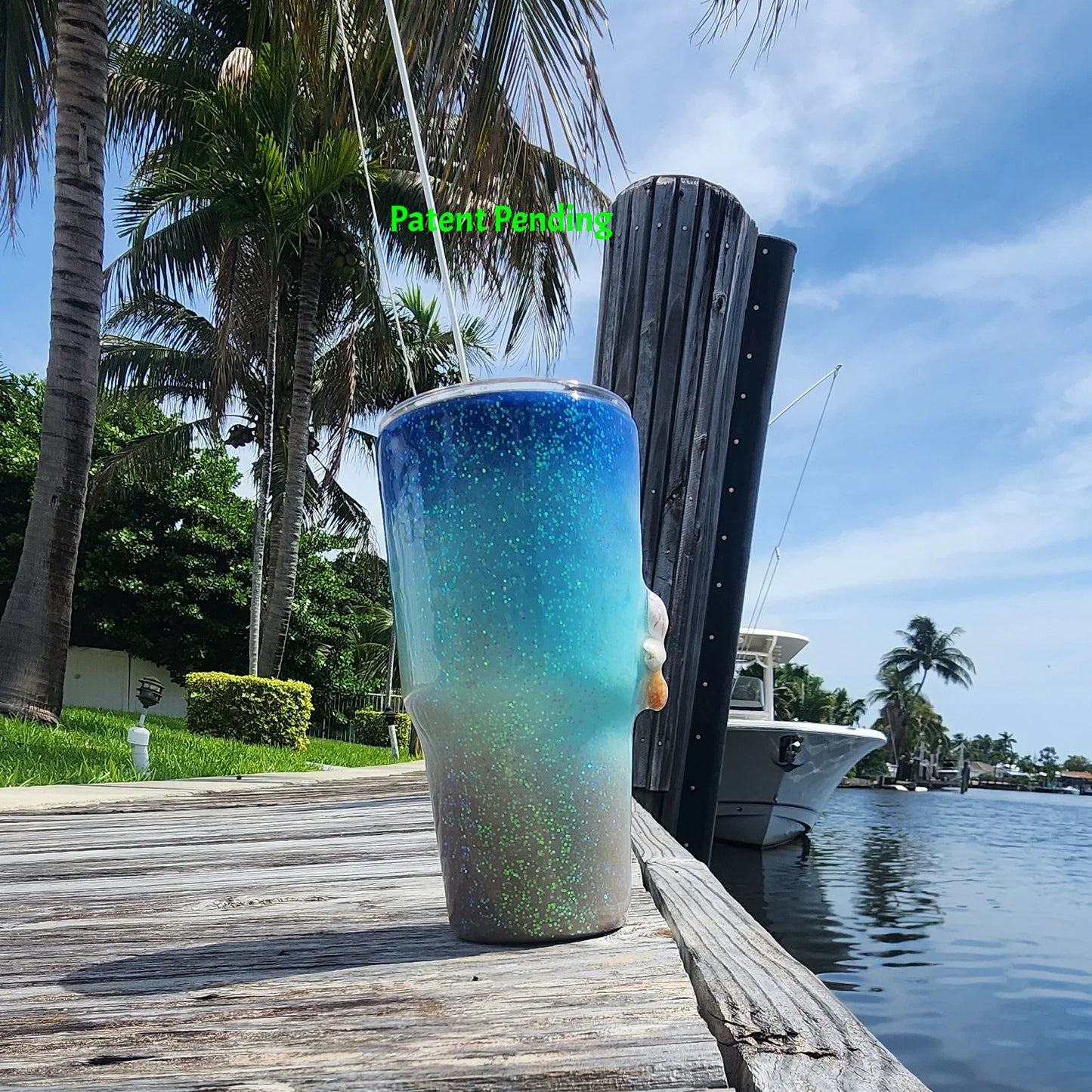 Left side view of Caribbean Glow Shell Glitter 30oz tumbler on South Florida marina dock showing holographic iridescent glitter color shift with palm trees and boat in background. Saltwater Sips™ by SoFlo Boho Boutique, Patent Pending.