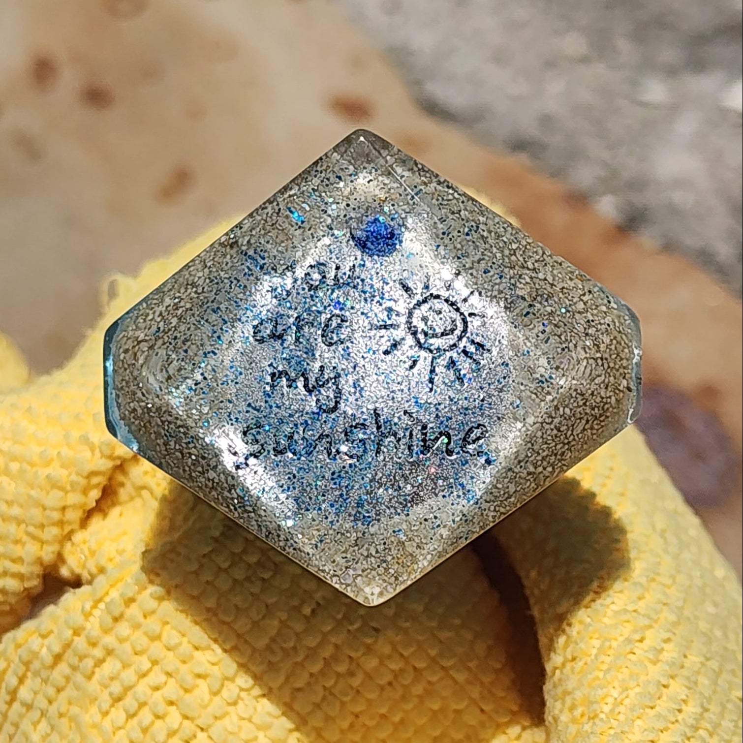 Close-up view of a geometric resin bottle stopper top, showcasing embedded beach sand, fine glitter, and the handwritten words You are my sunshine next to a small sketched sun.