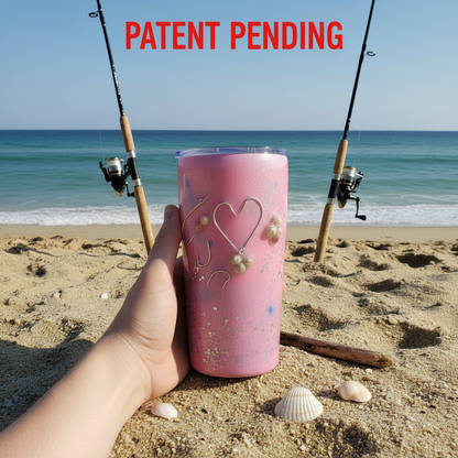 Pink tumbler with fish hook and seshells held on a beach with fishing rods and ocean in the background.