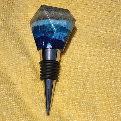 Handmade tapered geometric resin wine bottle stopper featuring a layered design of dark ocean blue, lighter turquoise waves, and a natural sand top on a shiny metal alloy base.