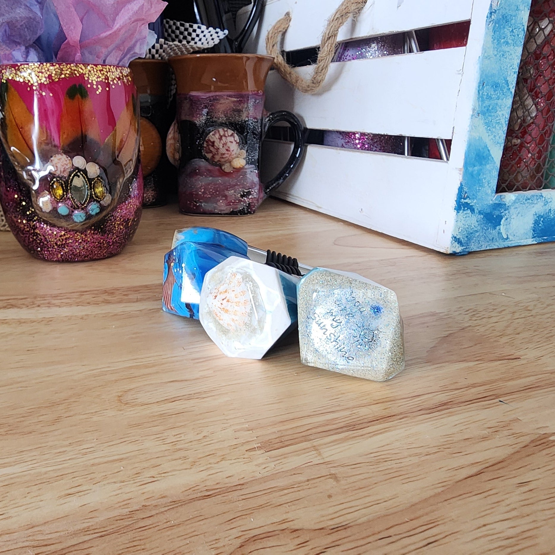 A cluster of handmade coastal resin bottle stoppers resting on a wooden countertop, clearly displaying a white-rimmed shell design and a sandy piece inscribed with "You are my sunshine" right in the foreground.