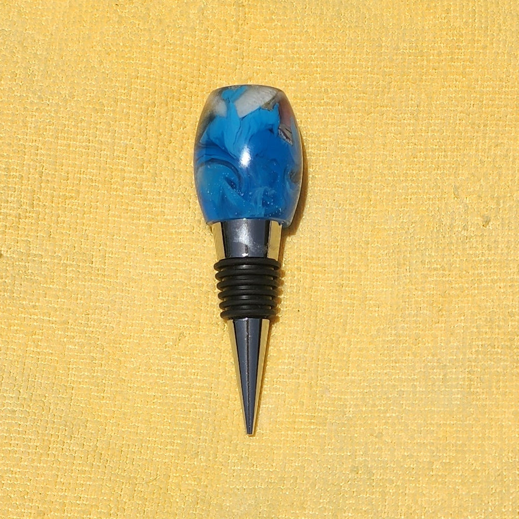 Close-up view of the rounded top of a resin bottle stopper, showcasing embedded real beach sand, a tiny white seashell, and swirling blue water effects.