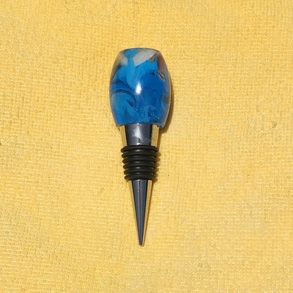 Close-up view of the rounded top of a resin bottle stopper, showcasing embedded real beach sand, a tiny white seashell, and swirling blue water effects.