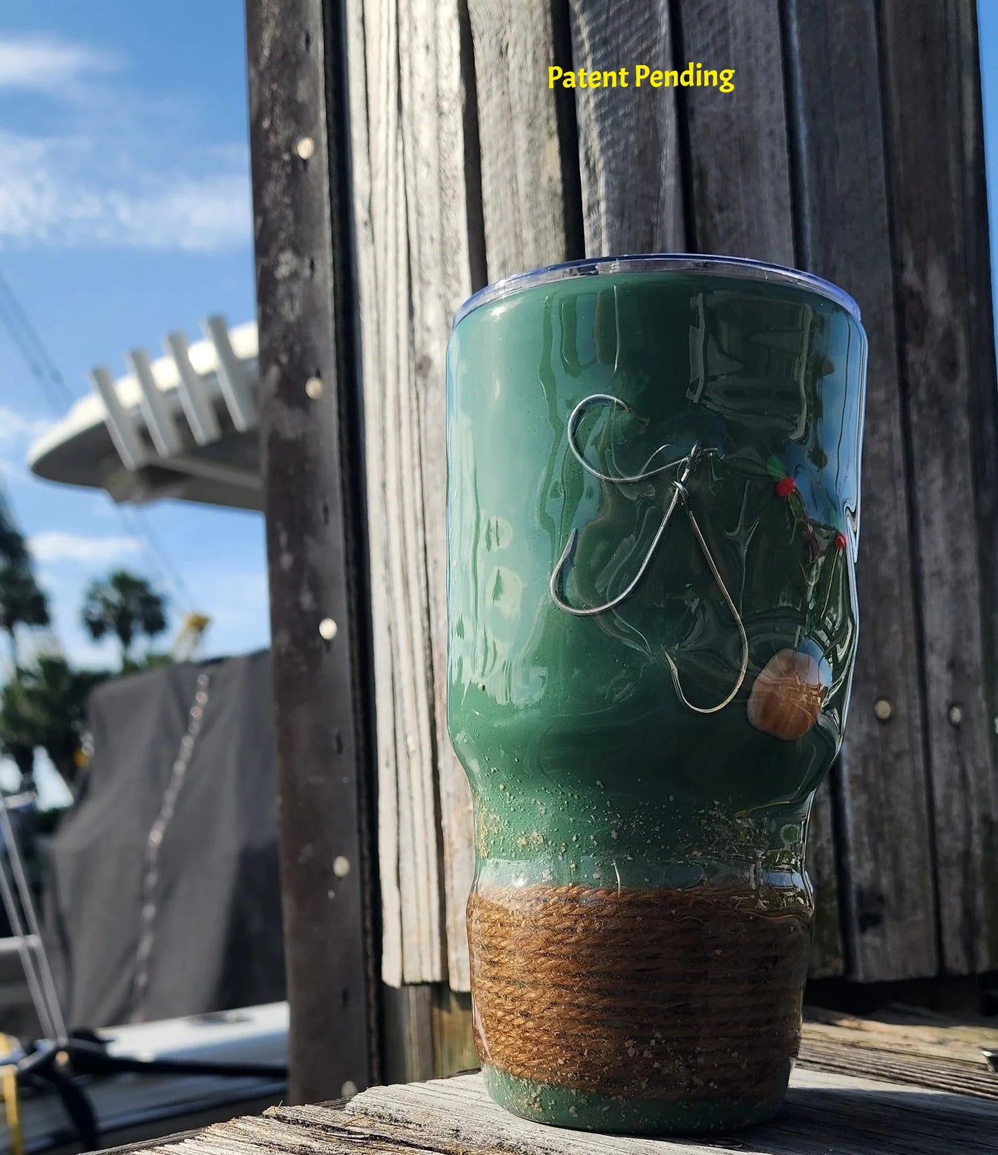 Marsh Tide 30oz Hook & Tumble Patent Pending fishing tumbler on marina dock showing real fish hooks, glow in the dark shrimp hooks and seashell with natural rope wrapped base against blue sky