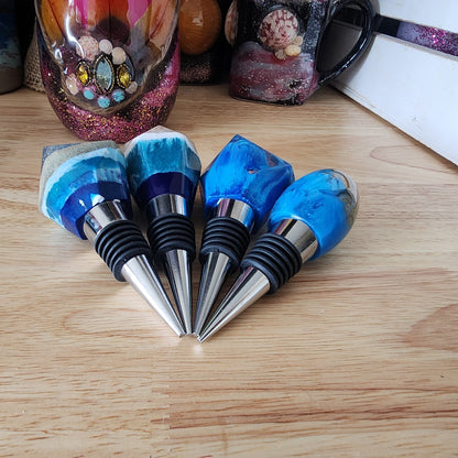 A close-up, front-facing view of four unique ocean-inspired resin wine bottle stoppers fanned out on a wooden table, showcasing their polished metal alloy bases, black rubber seals, and distinct layered beach and wave designs.