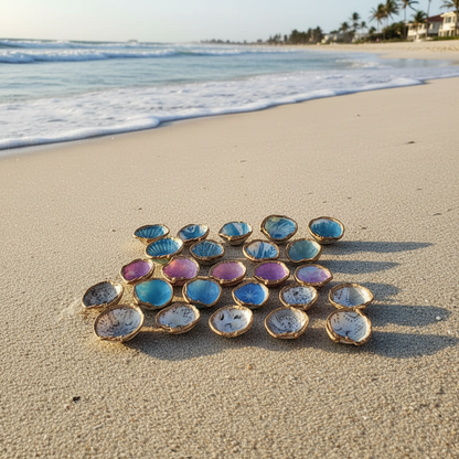 Handmade seashell fridge magnets arranged on the sand at the shoreline, gold-edged Lucina shells in blue, purple, and white patterns with ocean waves and beach houses in the background