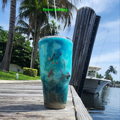 Handmade 30oz teal Caribbean coastal fishing tumbler with real fish hooks, glow-in-the-dark shrimp lures, silver crab and starfish charms, and sea snail shells - Hook & Tumble™ by SoFlo Boho Boutique, displayed on a South Florida marina dock with palm trees and water in the background.