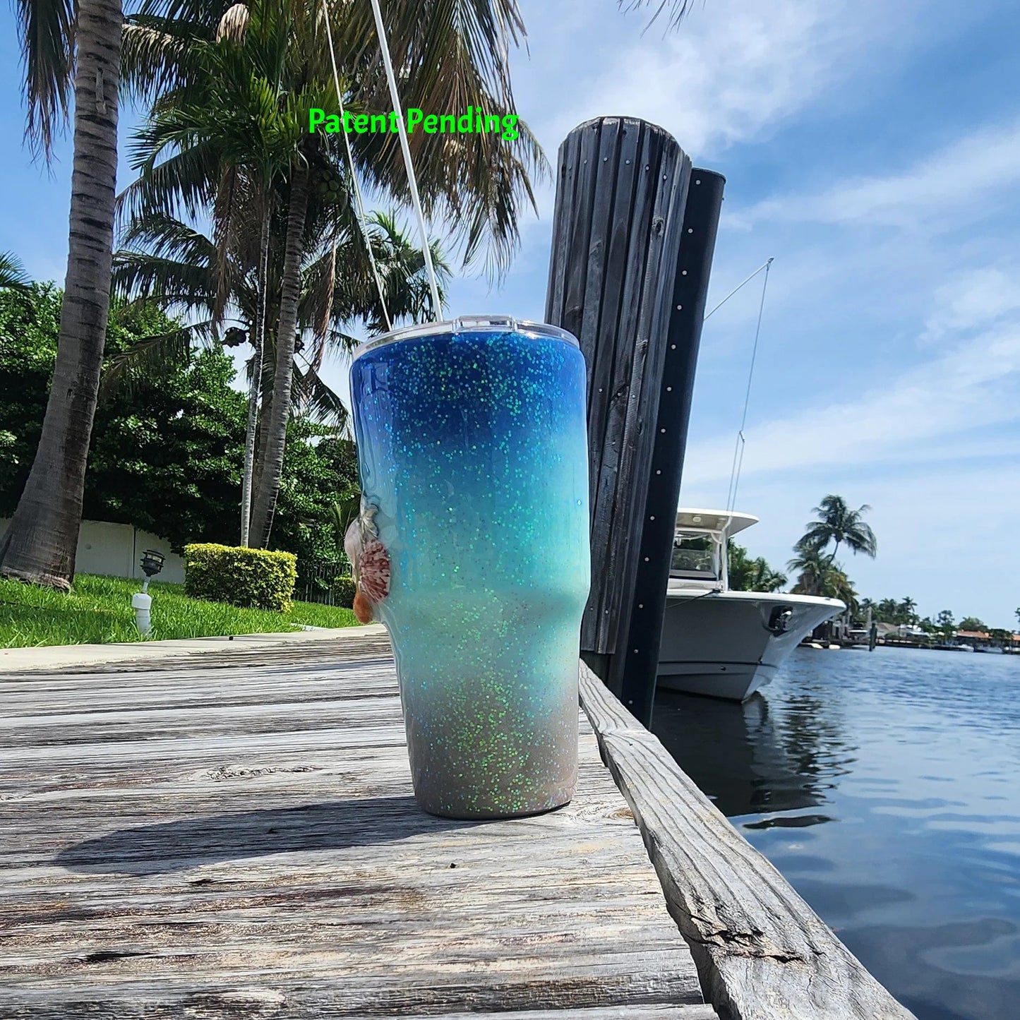 Right side view of Caribbean Glow Shell Glitter 30oz tumbler on South Florida marina dock showing real seashell and silver starfish charm detail with palm trees and boat in background. Saltwater Sips™ by SoFlo Boho Boutique, Patent Pending.